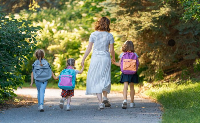Parent and pupils of primary school go hand in hand. Woman and girls with backpacks behind backs. Beginning of lessons. First day of fall.