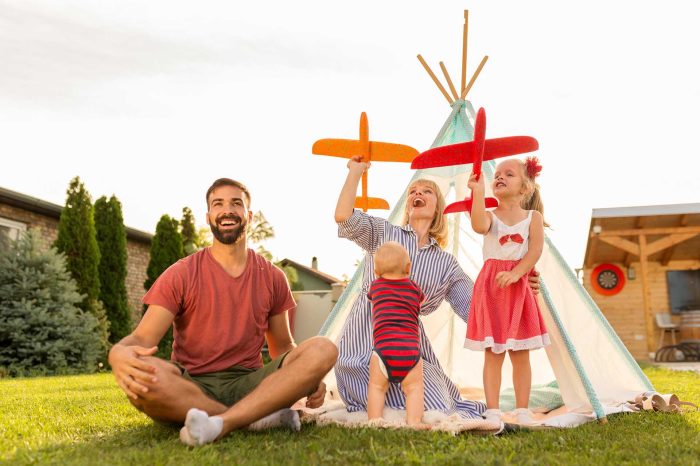 Beautiful young happy family having fun spending sunny summer day camping in the backyard, parents playing with their children, throwing toy airplanes